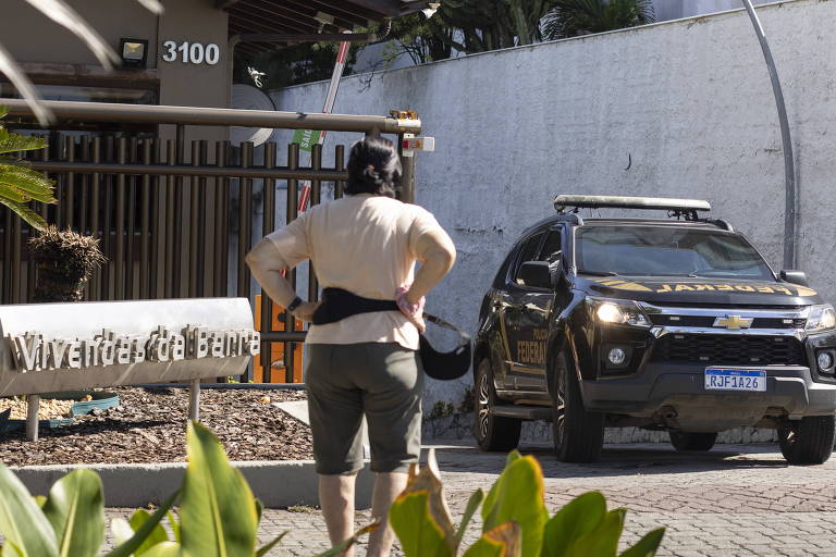 Mulher de costas assiste carro da PF saindo de garagem de condomínio
