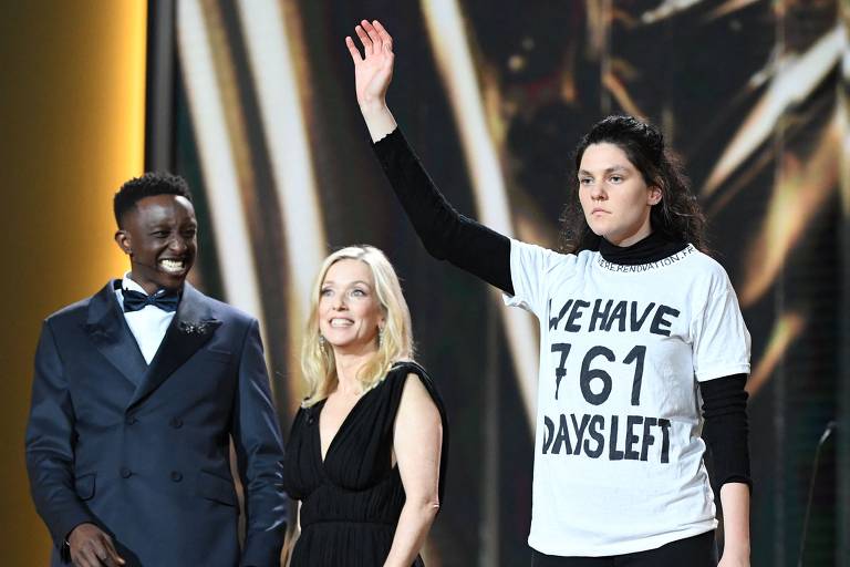 Mulher com a mão erguida ao lado dos dois apresentadores do prêmio