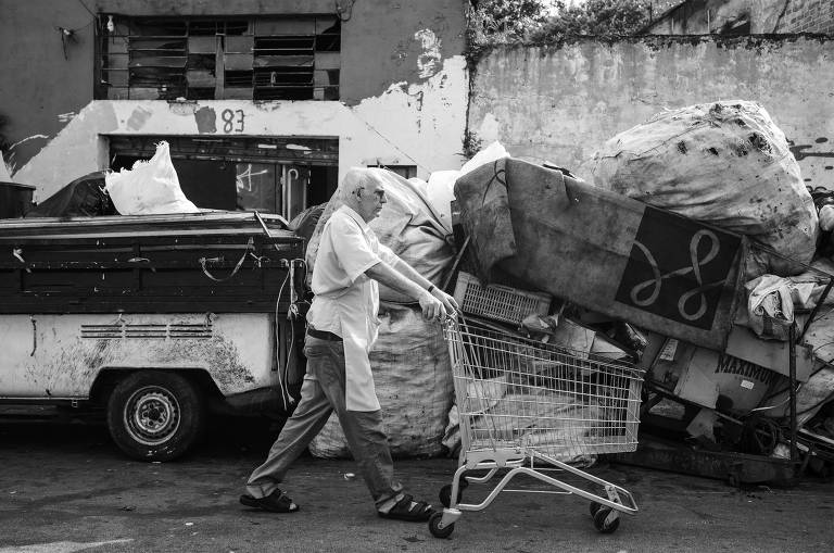 A imagem em preto e branco o homem empurra um carrinho de supermercado diante em uma rua com muita sucata acumulada na calçada