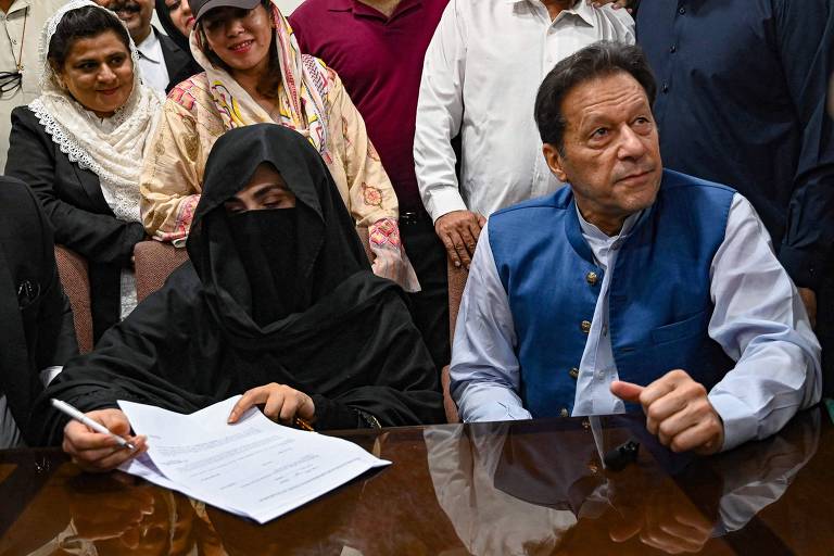Homem vestindo camisa e colete está sentado ao lado de mulher de vestimenta islâmica preta que só deixa os olhos de fora, com pessoas ao fundo