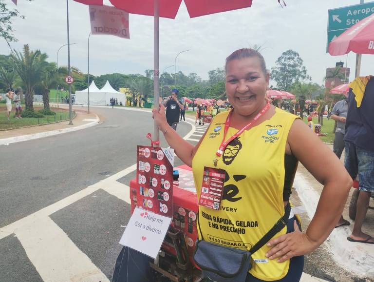ana paula posa exibindo carrinho de bebidas. que tem placa com texto "help me get a divorce", inglês para "ajude-me a conseguir um divórcio""
