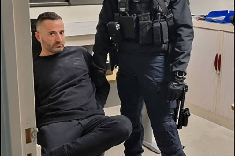 Homem branco de meia idade, sentado, com barba por fazer e colete anti bala está ao lado de policial com o rosto coberto 