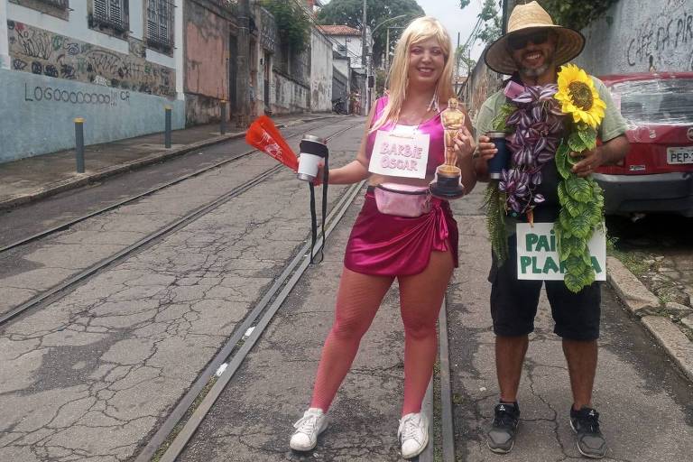 O casal Ricardo Pantoja, 48, e Aline Pantoja, 36, aproveita bloco Céu na Terra no bairro de Santa Teresa, no Rio de Janeiro, neste sábado (3). Ele segura um cartaz com a mensagem "pai de planta", e ela, um cartaz com a mensagem "Barbie Oscar