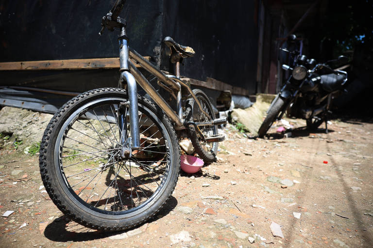 Bicicleta velha numa rua de terra, apoiada na parede de um barraco de madeira; ao fundo, uma motocicleta