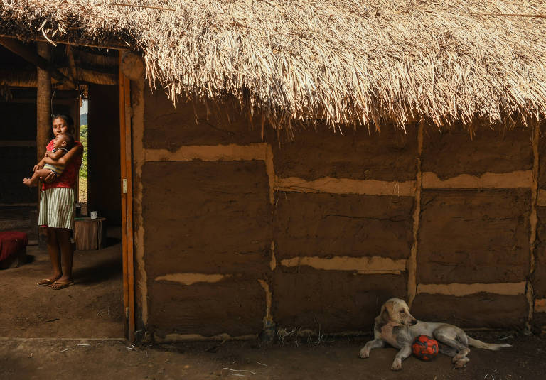 maysa segura o filho na porta de casa, uma construção simples, com teto de palha, e há um cachorro que descansa deitado no chão sob a sombra