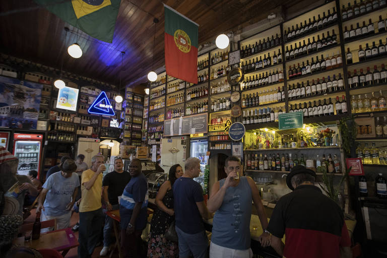 clientes em bar do rio com parede com prateleiras de garrafas até o teto e bandeiras de Brasil e Portugal 