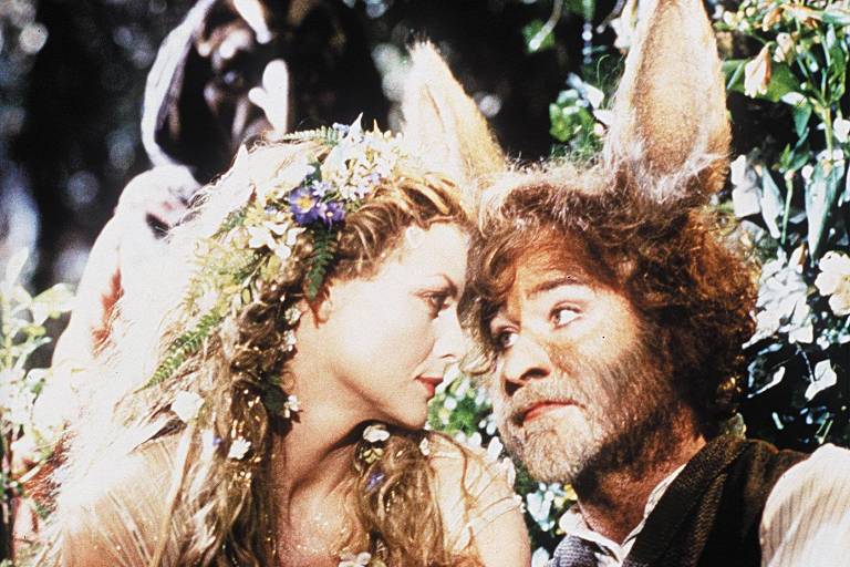 Uma mulher loira, com flores no cabelo, troca olhares com um homem barbudo com orelhas de burro