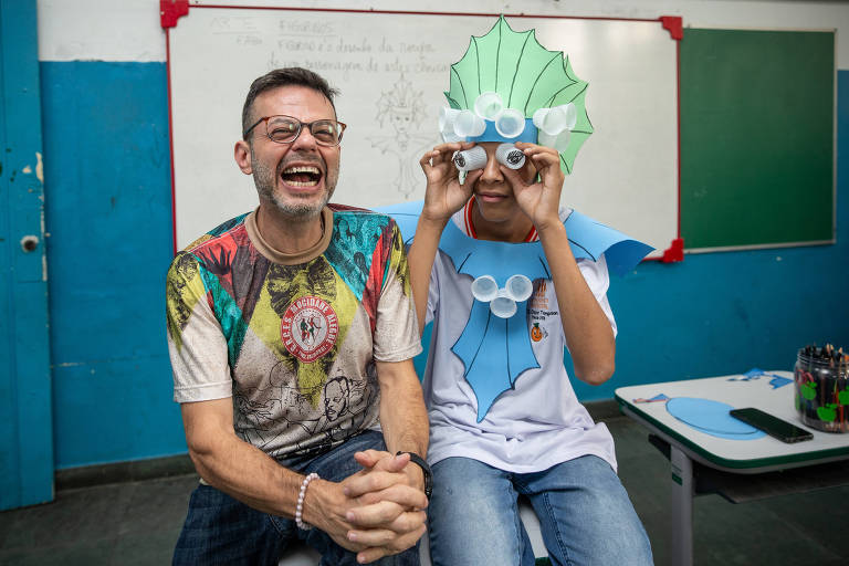 Homem sorridente está sentado em uma carteira de sala de aula ao lado de um adolescente que segura dois copos de plástico nos olhos e veste roupa de Carnaval improvisada com cartulina