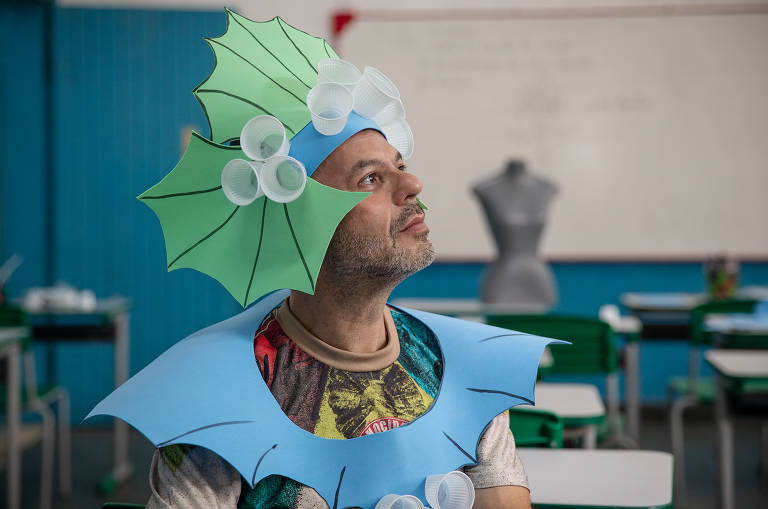 Homem branco está sentado em uma sala de aula, vestindo um cocar verde feito de cartolina verde e um colar de cartolina azul