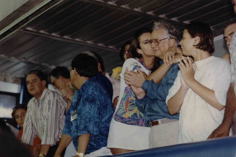 Mulher jovem de camiseta na altura da coxa cola rosto no de homem grisalho de camisa azul e calça cáqui
