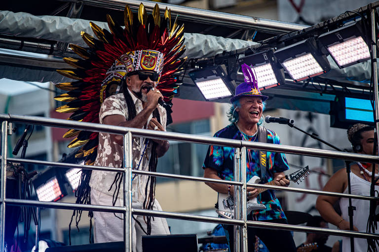 A imagem mostra uma cena de um evento musical. Dois homens vestidos com trajes típicos de povos indígenas estão em um palco， um deles usando um grande cocar de penas coloridas. Eles estão se apresentando para uma multidão. Há diversos equipamentos de iluminação e som ao redor deles. 