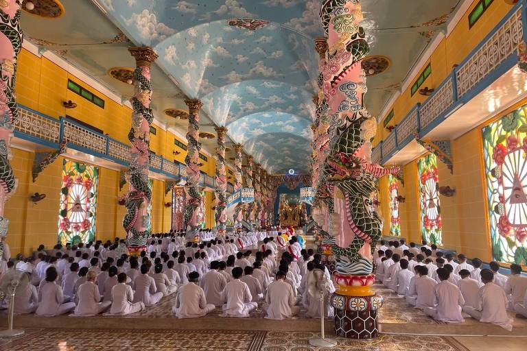 Pessoas sentadas no chão e enfileiradas no interior de um templo de teto alto e paredes decoradas