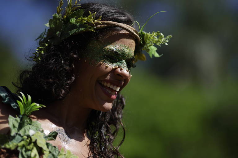 Close de uma foliona fantasiada e sorrindo; ela usa adereços de plantas e tem uma faixa pintada na altura dos olhos verde.