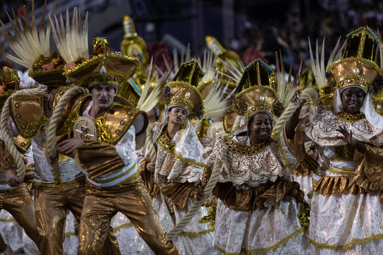 Imagem mostra integrantes de escola de samba dançando durante desfile. O grupo veste fantasias douradas e brancas. Ao fundo, o público assiste ao desfile na arquibancada, que aparece em desfoque na imagem 