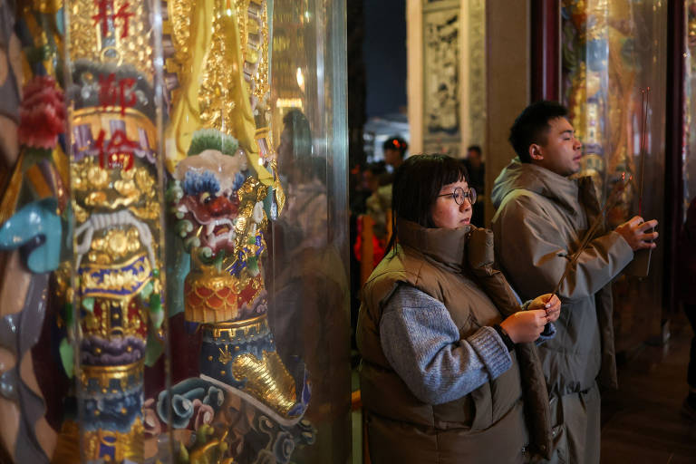 Homem e mulher de aparência asiática, vestindo casacos bege, estão de pé em frente a templo, rezando