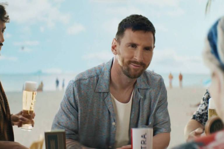 Messi está apoiado na bancada de um bar na praia. Ao lado dele, um homem segura uma cerveja