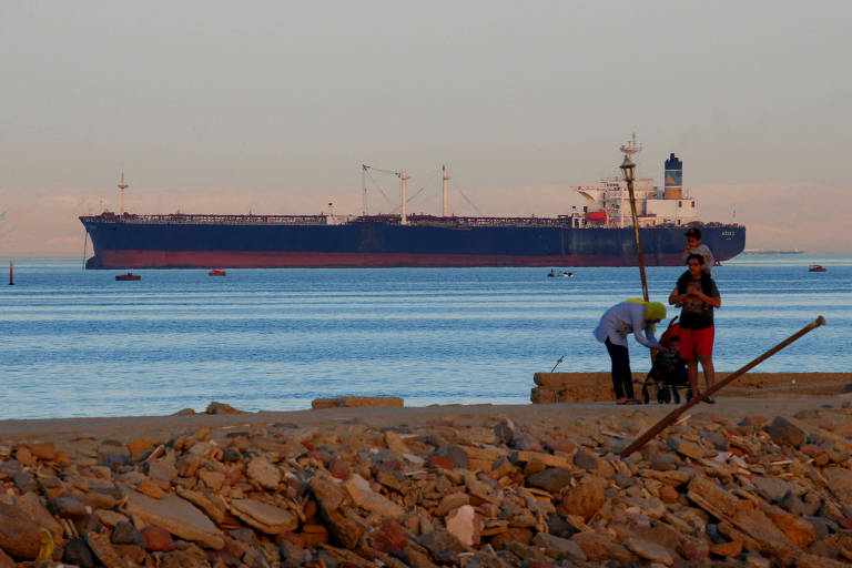 Imagem mostra navio cruzando golfo. Em primeiro plano, duas pessoas estão em uma praia cheia de pedras