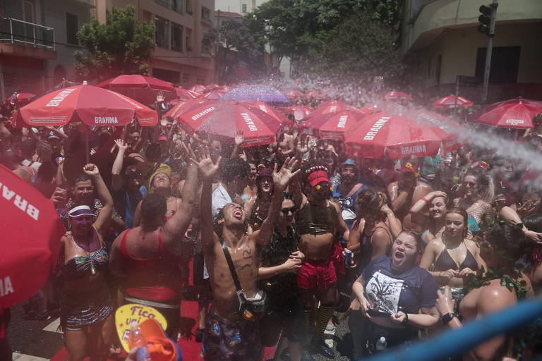 Centenas de foliões festejam o Carnaval em uma rua; água está sendo jogada sobre eles com uma mangueira