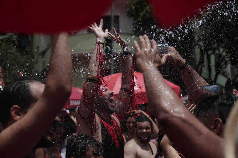 A imagem mostra um grupo de pessoas se divertindo em uma festa ao ar livre. Elas estão levantando as mãos e se molhando com água， enquanto algumas seguram copos. 