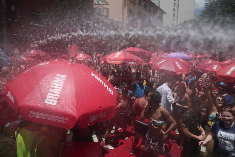 A imagem mostra uma grande multidão reunida em um evento ao ar livre， com várias pessoas sob guarda-sóis vermelhos. Há um jato de água sendo pulverizado sobre os participantes， que parecem se divertir