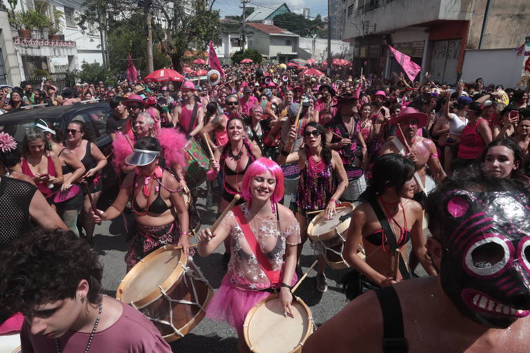 A imagem mostra uma grande multidão participando de um desfile de carnaval. As pessoas estão vestidas com roupas predominantemente rosa e preto， algumas usando fantasias e acessórios coloridos. Há pessoas tocando tambores e dançando， enquanto outras seguram bandeiras.