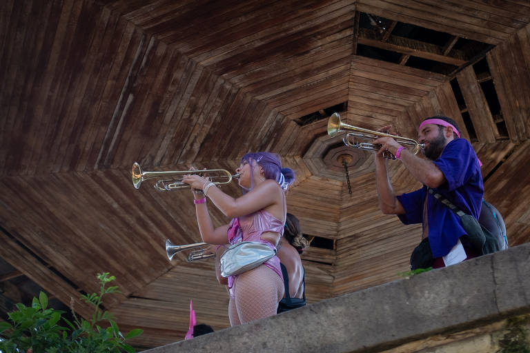 A imagem mostra dois músicos em uma apresentação. Eles estão em uma estrutura elevada， tocando trompetes. A mulher à esquerda tem cabelo roxo e usa uma roupa colorida， enquanto o homem à direita está vestido com uma camisa azul e um lenço na cabeça. O fundo é uma construção de madeira com um teto hexagonal.
