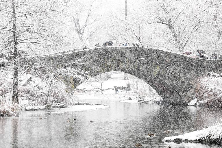 Foto mostra uma ponte sobre um rio com neve caindo e árvores sem folhas