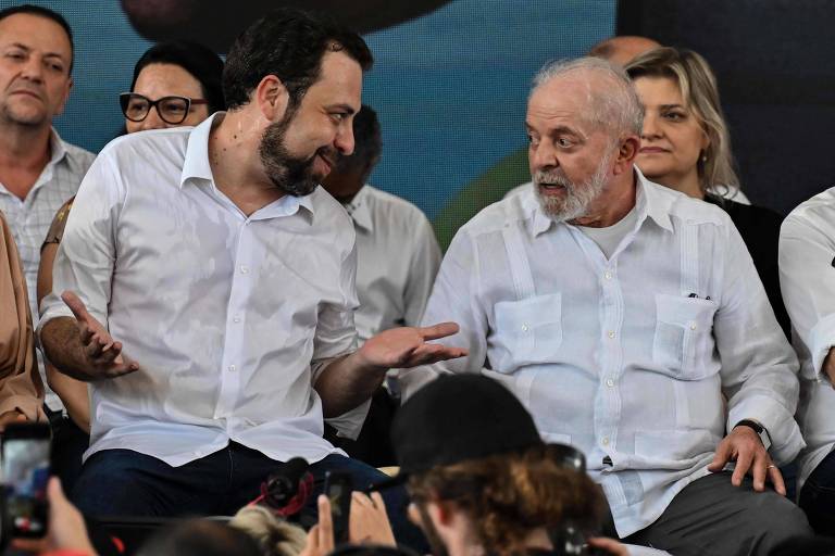 Foto mostra Boulos e Lula, sentados em cadeiras no palco. Ambos vestem camisas brancas e estão conversando