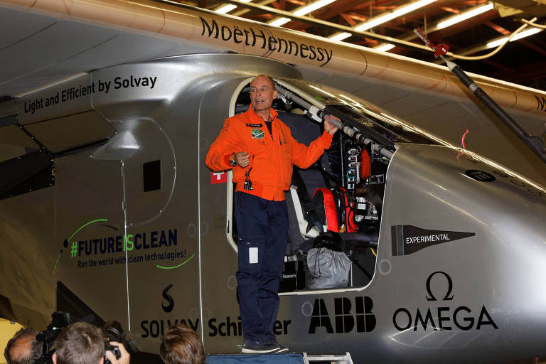 Bertrand Piccard é um homem branco que veste um casaco laranja e calça escura. Ele está ao lado de um avião.