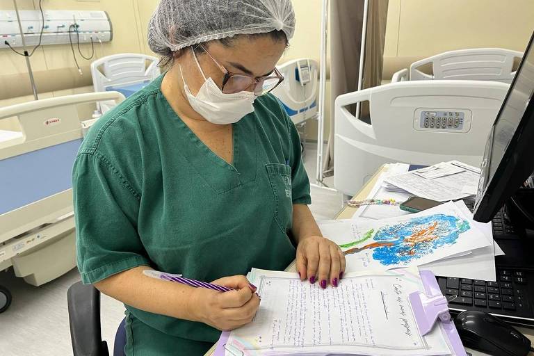Mulher com roupa verde, máscara e touca brancas, escreve carta dentro de um hospital