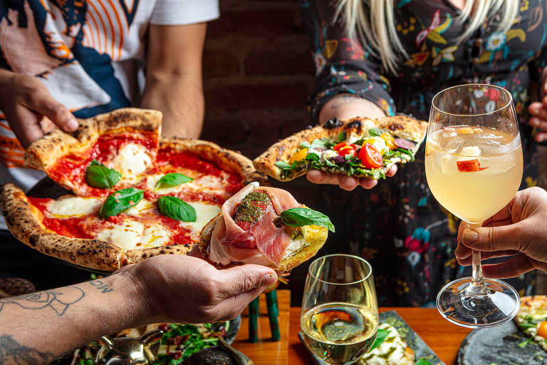 A foto mostra mãos segurando pizzas e taças de drinques