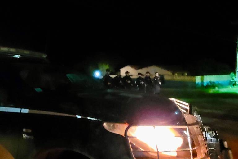 Carro de polícia com farol ligado, à noite, com policiais ao fundo