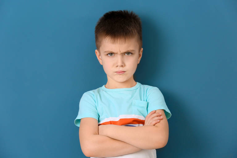Menino branco bravo, nervoso de braços cruzados, usando camiseta azul de listra vermelha e branca, atrás, fundo azul