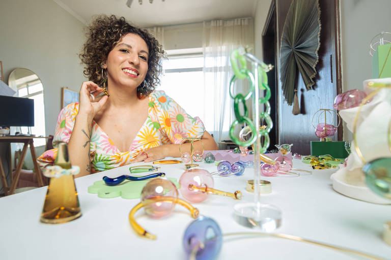 A foto mostra a designer Mariana Genoveze, uma mulher de cabelos cacheados, curtos, com um vestido florido. Na sua frente, as peças que produz em seu ateliê, feitas de vidro.