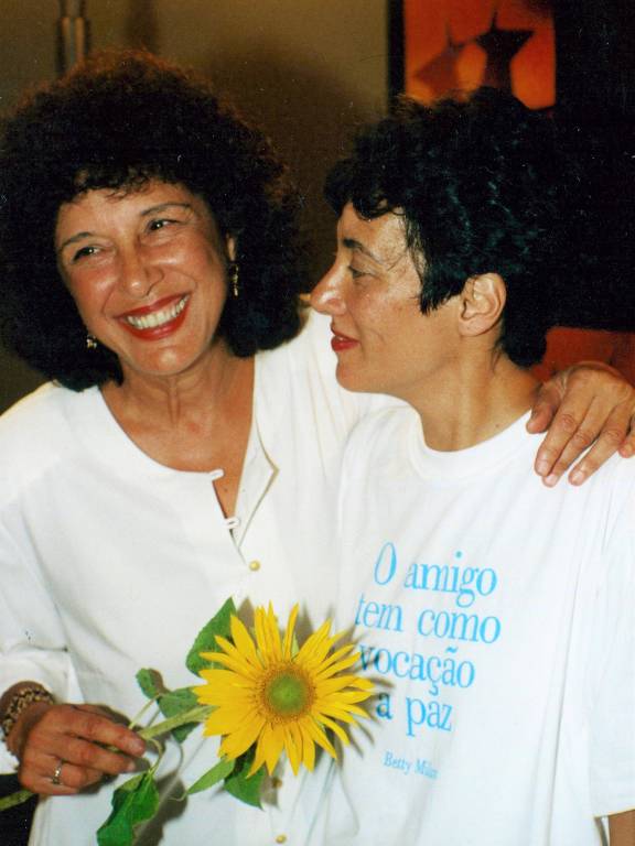 duas mulheres brancas se abraçam; uma delas segura uma flor amarela