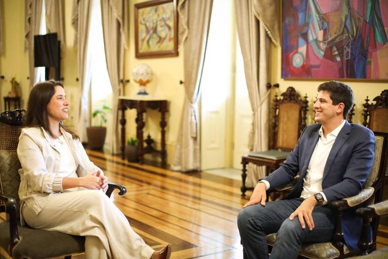 Raquel Lyra sentada vestida de branco olha para o prefeito João Campos, que também está em uma cadeira com terno azul e camisa branca. Ambos sorriem e estão em uma sala fechada