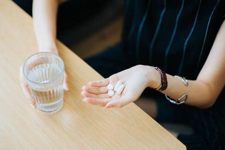 Foto mostra os braços e mãos de uma mulher branca que segura comprimidos e um copo d'água