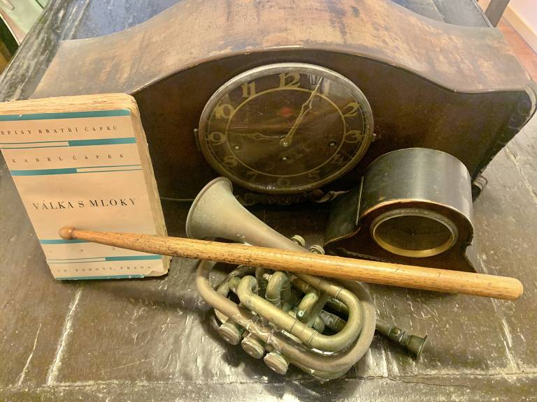 Foto mostra objetos antigos em uma mesa: relógios de mesa, um cornet (trompete) americano, uma baqueta de bateria e a primeira edição, em tcheco, do livro 'A Guerra das Salamandras'
