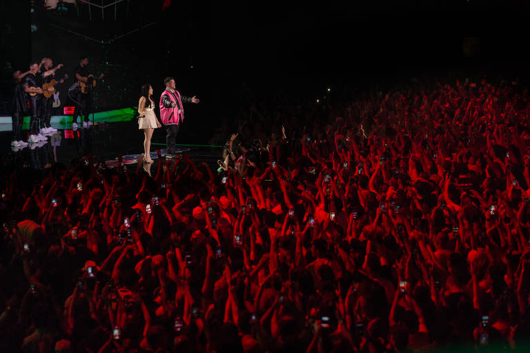 Show do cantor Ferrugem, que veste um colete rosa e calça preta, com participação de Mari Fernandez, que usa um vestido claro acima do joelho, no palco na Vibra SP. Ao fundo está a banda. A plateia é iluminada com luzes vermelhas