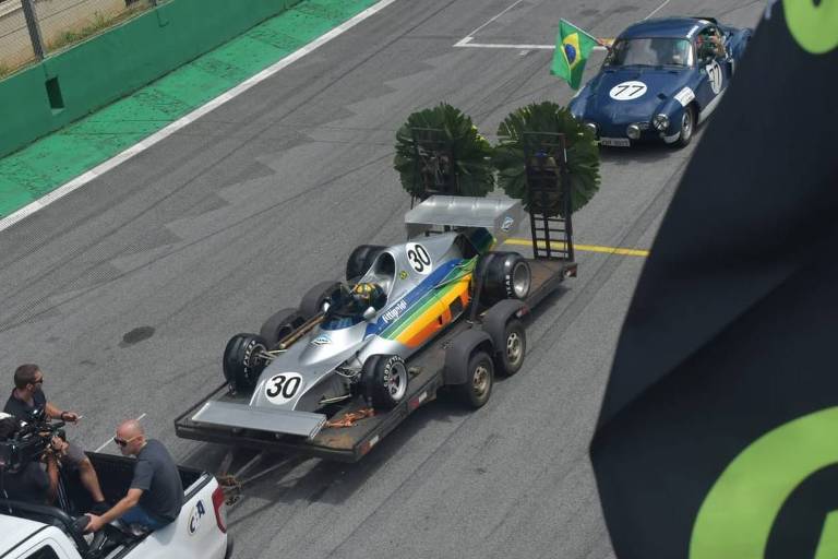 Imagem mostra um carro de corrida antigo sendo rebocado em cima de uma carretinha ao longo de uma pista de automobilismo.