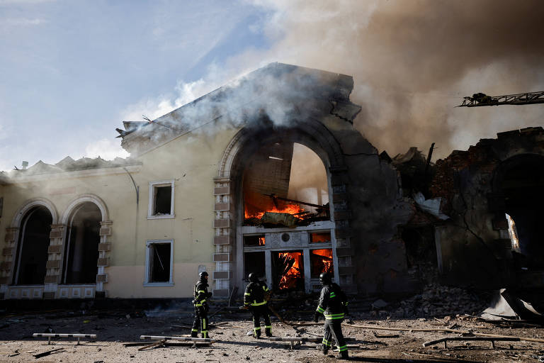 A fachada de uma construção está pegando fogo e três pessoas vestidas com roupas de proteção usam mangueiras para combater as chamas