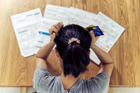Vista superior de uma jovem asiática estressada com as mãos segurando a cabeça preocupada em encontrar dinheiro para pagar dívidas de cartão de crédito e todas as contas de empréstimo. Conceito de problema financeiro. (Foto: Kawee/Adobe Stock )