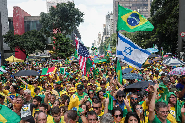 Imagem de uma grande manifestação em uma avenida, com muitas pessoas vestindo roupas nas cores verde e amarelo. Diversas bandeiras são visíveis, incluindo as do Brasil, Estados Unidos, Israel e outras. A manifestação ocorre em um ambiente urbano com prédios altos ao fundo.