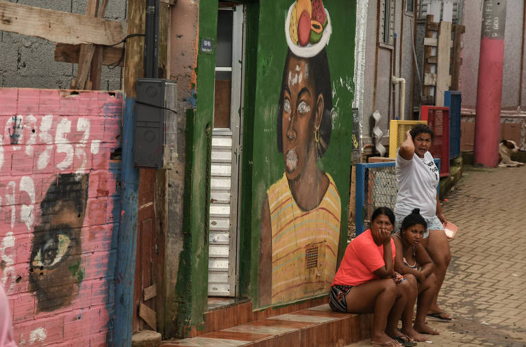 Imagem mostra duas moradoras sentadas em uma calçada e outra moradora em pé. Elas olham para a câmera de longe. As paredes das casas que aparecem ao fundo são pintadas com murais coloridos