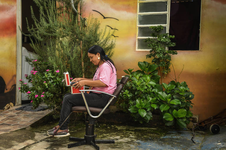 Imagem mostra Jaqueline Silva, de lado, sentada na cadeira usada para fazer design de sobrancelhas. Ela segura uma caixa vermelha. O ambiente tem plantas e uma parede colorida de tons amarelos e laranjas com uma janela e uma porta mais ao fundo e à esquerda 