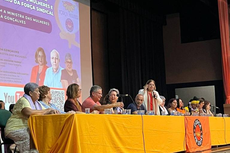 mulher branca em pé, com microfone, atrás de mesa com várias pessoas sentadas