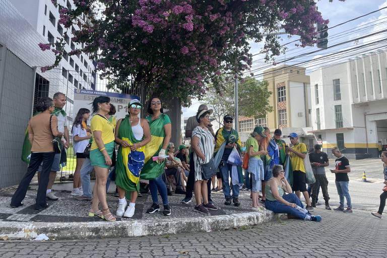 Pessoas vestidas de verde e amarelo reunidas em calçada em rua sob sol