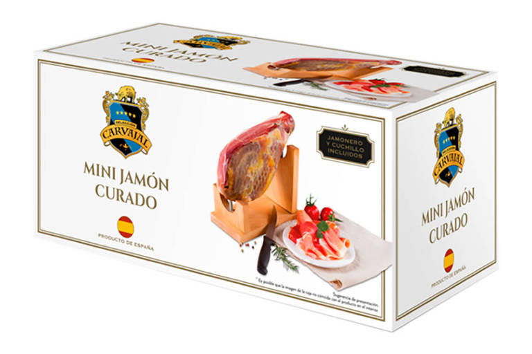 Na foto, uma caixa branco com imagens de um mini Jamon apoiado sobre um suporte de madeira, e um prato branco com fatias de Jamon. 