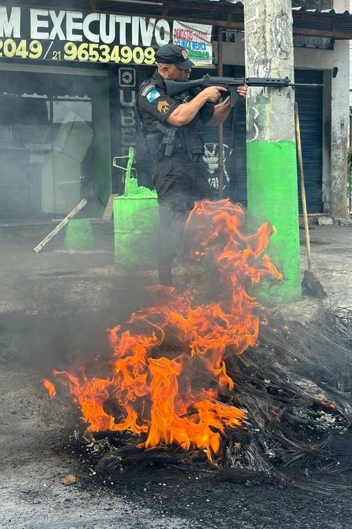 Policial aponta fuzil atrás de poste ao lado de uma barricada com óleo e fogo