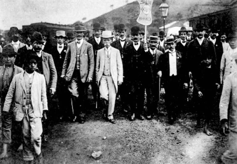 foto preto e banca de jurista Ruy Barbosa de cartola e terno brancos cercado de homens de terno